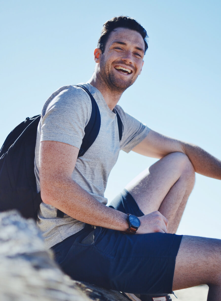 Smiling man with backpack sitting on a rock under clear blue sky.