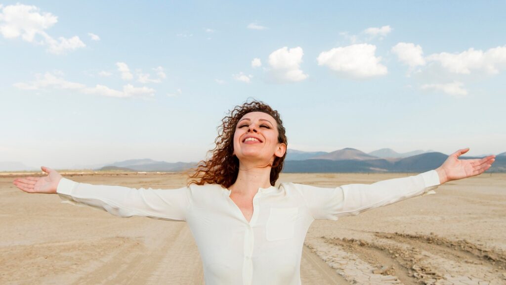 Joyful woman in white shirt standing arms open in desert-like area.