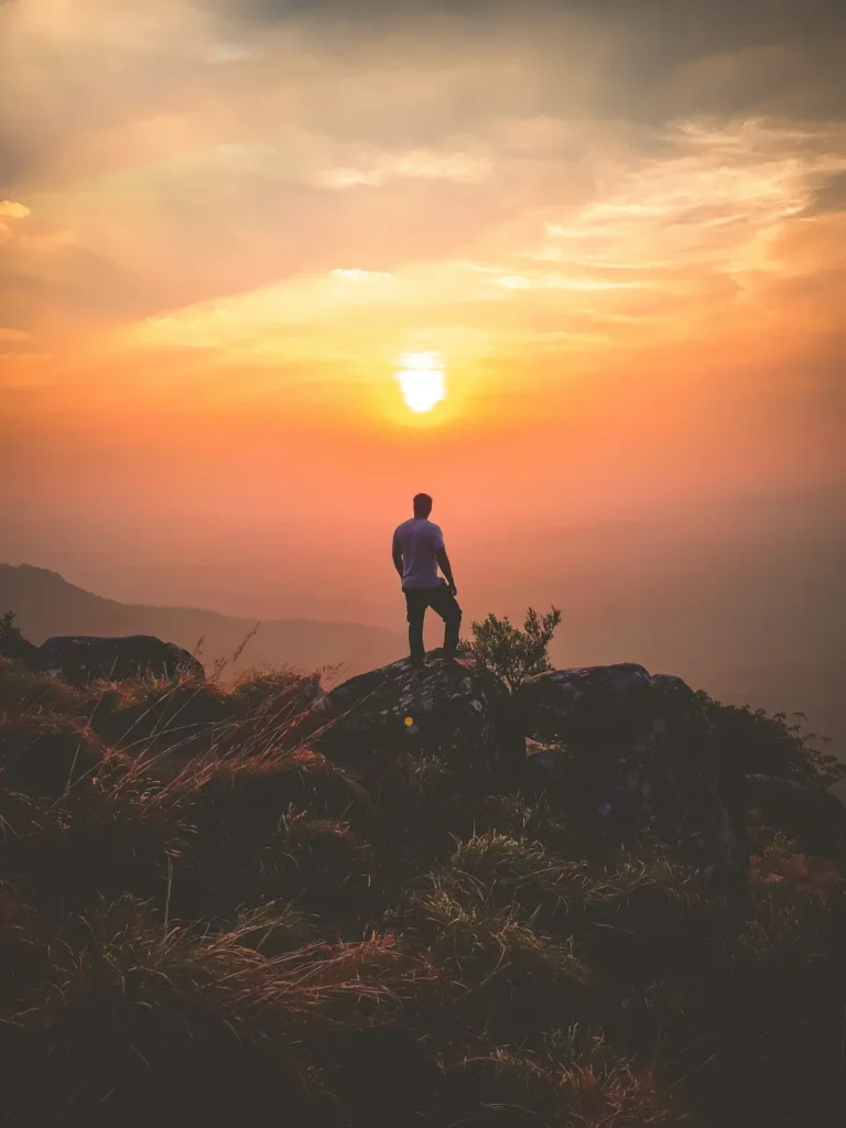 Man standing on a cliff watching sunset over mountain landscape.