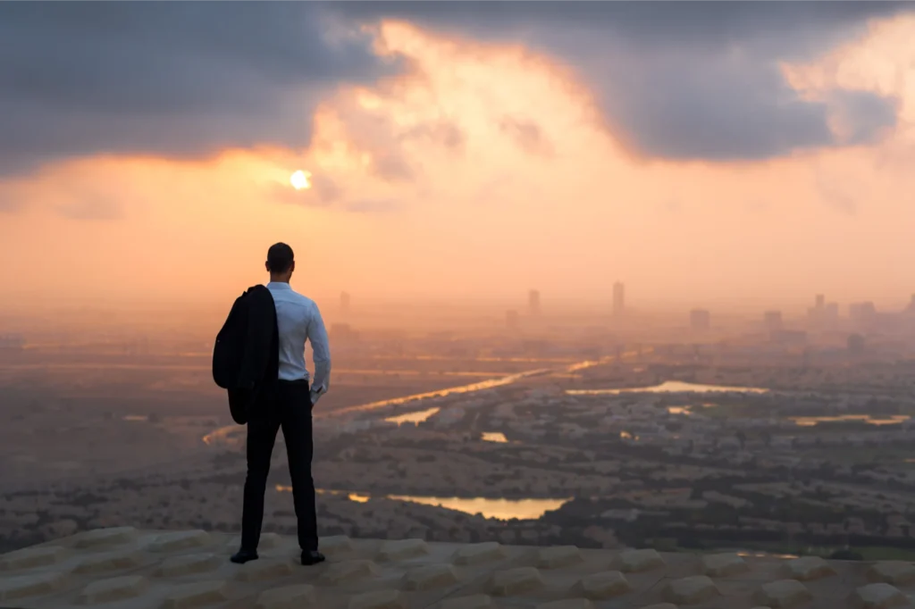 Businessman standing on a high viewpoint overlooking a city at sunrise.